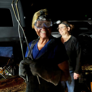 Two smiling potters with headlamps stand outdoors at night, one in safety goggles and heavy gloves holding long tongs, with The Clay Hole, a pottery studio in Draper, Utah
