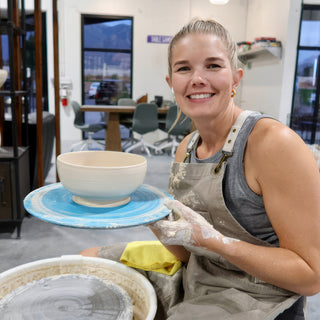 A smiling potter with clay-covered hands holds a pale bowl on a blue disk beside a pottery wheel at The Clay Hole in Draper, Utah