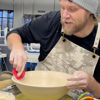 A potter smoothing the rim of a wide, shallow clay bowl with a red sponge while steadying the side at The Clay Hole in Draper, Utah