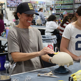 A potter in a black cap smiling while smoothing a pale unglazed dome-shaped piece with a yellow tool at The Clay Hole in Draper, Utah