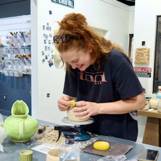 A potter laughs while smoothing a clay piece on a turntable with a yellow sponge, beside a green glazed vessel at The Clay Hole in Draper, Utah