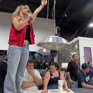 A person in a red vest speaks into a microphone with one arm raised above a seated crowd at The Clay Hole in Draper, Utah