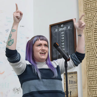 A person with purple hair speaking into a microphone with both arms raised and one finger pointing up at The Clay Hole in Draper, Utah