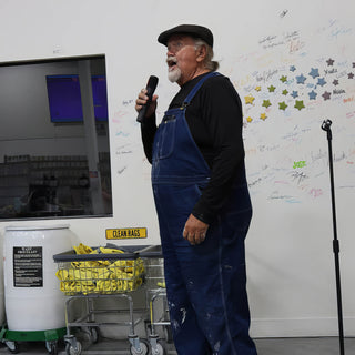 A person in a flat cap and overalls speaking into a microphone in front of a wall of signatures at The Clay Hole in Draper, Utah