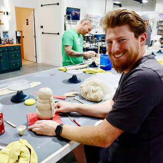 A smiling potter shapes a stacked pale clay form with a handle on a red board at a worktable at The Clay Hole in Draper, Utah