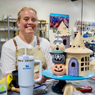 A smiling potter poses with a stacked pumpkin, cat and hatted face figure and a tan house with cutout windows at The Clay Hole in Draper, Utah