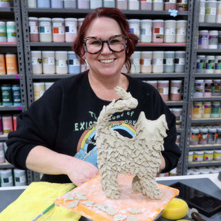 A smiling potter holds a standing clay animal covered in small layered clay tufts on an orange board at The Clay Hole in Draper, Utah