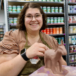 A smiling potter touches up a standing brown clay animal with tall pointed ears on a lavender board at The Clay Hole in Draper, Utah
