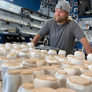 A potter in a gray beanie leans over a crowded table of white vessels with faceted shapes and tan tops at The Clay Hole in Draper, Utah