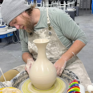 A potter in a gray beanie shaping a tall bottle-shaped vase with a flared rim on a pottery wheel at The Clay Hole in Draper, Utah