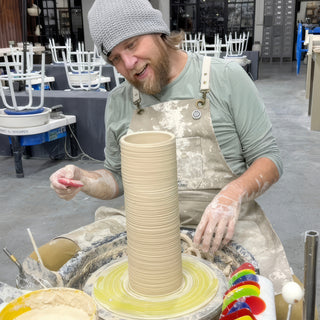 A potter holding a red sponge beside a very tall clay cylinder with ridged rings on a pottery wheel at The Clay Hole in Draper, Utah