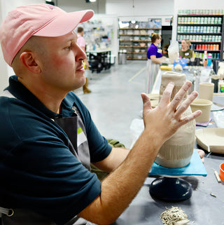 A potter in a pink cap holds a hand up beside a tall pale clay vase with fine vertical lines at The Clay Hole in Draper, Utah