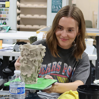 A smiling potter holds a tall pale clay vessel covered in sculpted flowers and leaves on a green board at The Clay Hole in Draper, Utah