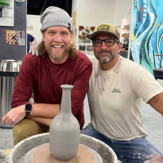 Two potters leaning in behind a tall pale gray bottle-shaped vase with a narrow neck on a pottery wheel at The Clay Hole in Draper, Utah