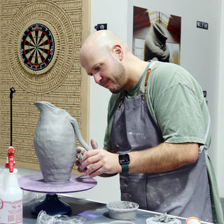 A potter works on the curved handle of a tall gray clay pitcher on a purple turntable at The Clay Hole in Draper, Utah