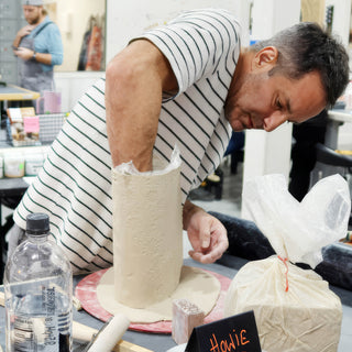 A potter with an arm inside a tall patterned clay cylinder sitting on a flat clay base at The Clay Hole in Draper, Utah