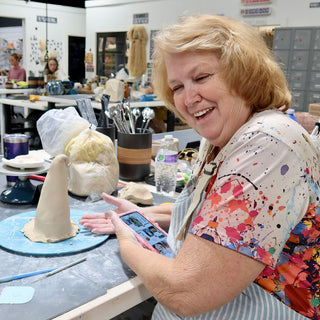 A laughing potter holds a phone beside a tall pointed clay form with a flared base on a blue board at The Clay Hole in Draper, Utah