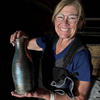 A smiling person in glasses holds a tall narrow vase with ridges and a shimmering blue and gray metallic finish at The Clay Hole in Draper, Utah