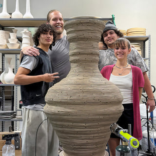 Four people pose beside a very tall unfired vase covered in fine horizontal ridges, one making a peace sign at The Clay Hole in Draper, Utah