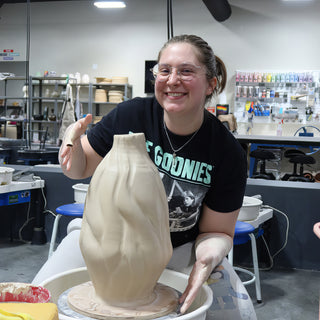 A smiling potter behind a tall tan vase with a twisted, ridged body sitting on a pottery wheel at The Clay Hole in Draper, Utah