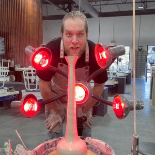 A potter with a wide-eyed look leaning over a tall slender pale vase on a wheel ringed by red lamps at The Clay Hole in Draper, Utah