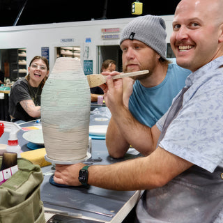 A smiling potter holds a wide brush beside a tall vase with pale green and tan bands, another potter behind at The Clay Hole in Draper, Utah