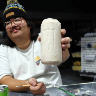 A potter in a beanie and glasses holds up a tall white cylinder with scratched surface marks at The Clay Hole in Draper, Utah