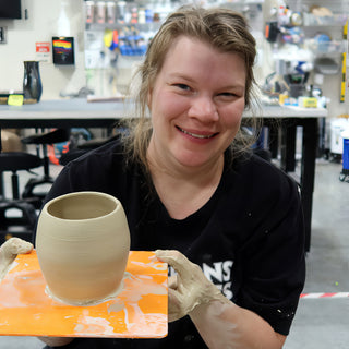 A smiling potter with muddy hands holds a tan rounded vessel with a wide opening on an orange board at The Clay Hole in Draper, Utah