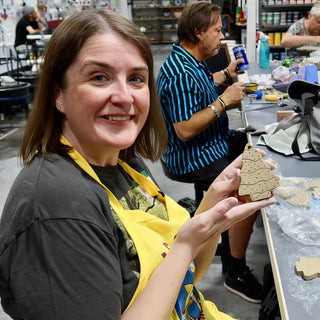 A potter in a yellow apron holds up a tan tree-shaped ornament with a textured surface at The Clay Hole in Draper, Utah