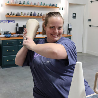 A smiling potter holding a tall, tapered clay piece up on one shoulder like a baseball bat at The Clay Hole in Draper, Utah