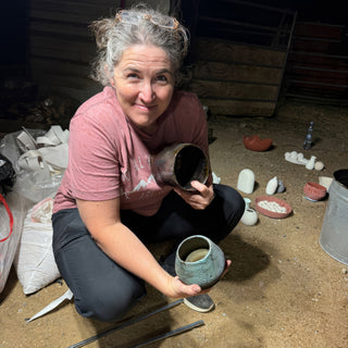 A potter kneeling outdoors at night holds a dark glossy vessel and a teal green cup, with The Clay Hole, a pottery studio in Draper, Utah