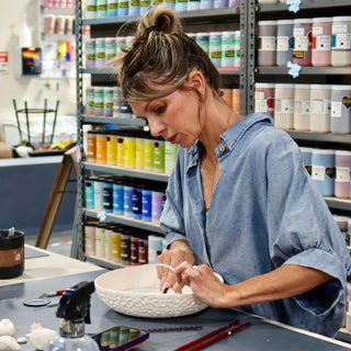 A potter in a blue shirt works inside a white bowl with a dimpled outside, glaze shelves behind, with The Clay Hole, a pottery studio in Draper, Utah