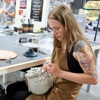 A potter with a tattooed arm works a fine tool over a pale vessel with wavy ridges and clustered shapes at The Clay Hole in Draper, Utah