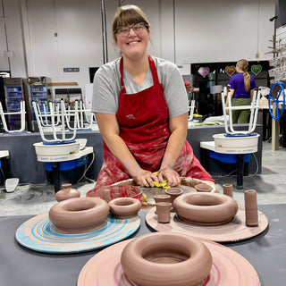 A smiling potter in a red apron sits behind thick brown clay rings and small cups on swirled boards at The Clay Hole in Draper, Utah