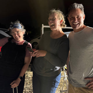 Three friends smile and pose close together outdoors at night, one in a dark apron with goggles on their head, with The Clay Hole, a pottery studio in Draper, Utah