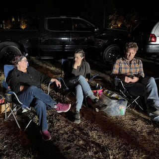 Three friends sit in camp chairs on dry grass at night with a black pickup truck parked behind them at The Clay Hole in Draper, Utah