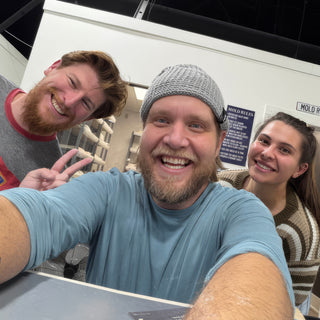 Three smiling people squeeze into a selfie, one flashing a peace sign, with shelves of white pieces behind at The Clay Hole in Draper, Utah
