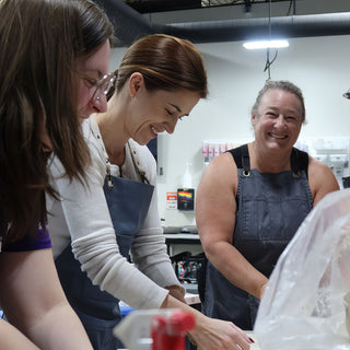 Two potters in aprons lean over their work at a long table while a third smiles at the camera at The Clay Hole in Draper, Utah