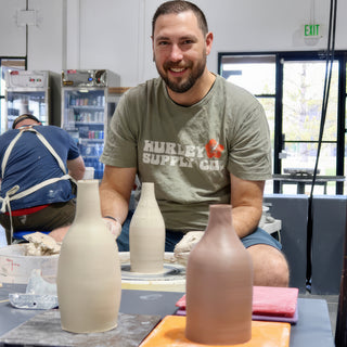 A smiling potter sits at a pottery wheel behind three tall bottle-shaped vases in gray and brown clay at The Clay Hole in Draper, Utah