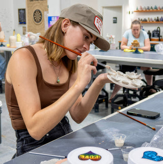 A potter in a tan cap works on tiny pale clay flower shapes on a plate using a thin brush at The Clay Hole in Draper, Utah