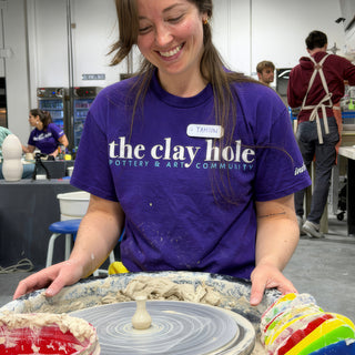 A smiling potter in a purple t-shirt resting hands on a pottery wheel with a tiny pale vase on it at The Clay Hole in Draper, Utah