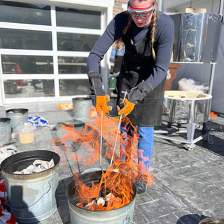A potter in goggles, heavy gloves and a black apron holds long tongs over a metal bucket full of flames at The Clay Hole in Draper, Utah