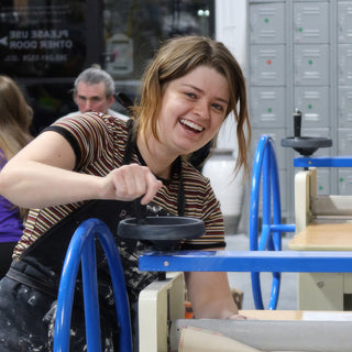 A smiling potter turning the blue hand wheel of a slab roller with metal lockers in the background at The Clay Hole in Draper, Utah