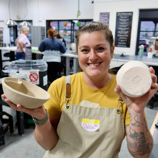 A smiling potter holding a rough tan bowl in one hand and a pale upturned bowl in the other at The Clay Hole in Draper, Utah
