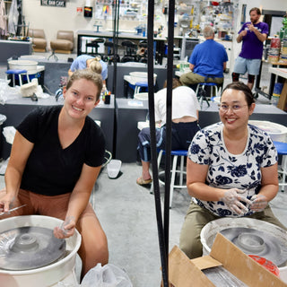Two smiling potters sit at wheels with gray clay on the wheel heads while others work in the busy room at The Clay Hole in Draper, Utah