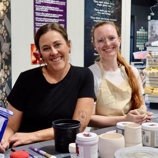 Two smiling potters sit at a table with unglazed cups, glaze jars and a red sponge in front of them at The Clay Hole in Draper, Utah