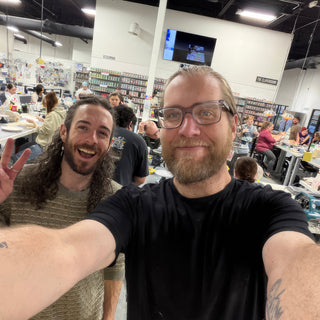 Two people take a selfie in a busy studio, one flashing a peace sign, with members working behind them at The Clay Hole in Draper, Utah