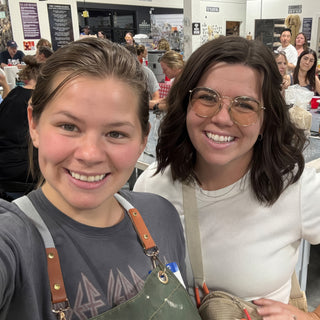 Two friends smile in a close selfie, one in a green apron, with a seated crowd behind them at The Clay Hole in Draper, Utah