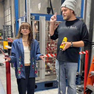 Two people stand in front of a tall blue metal rack, one holding a yellow tool at The Clay Hole in Draper, Utah