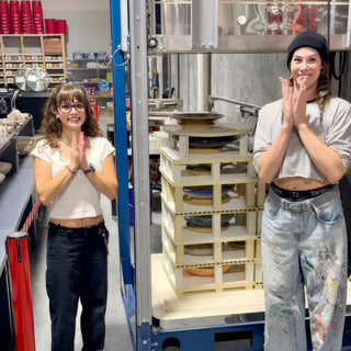 Two people smile and clap beside a tall stack of plates separated by shelf posts inside a blue metal frame at The Clay Hole in Draper, Utah